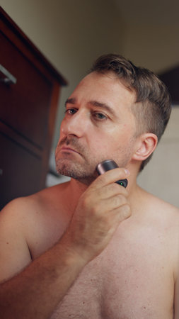 Vertical photo Individual reviews trimmed jawline under warm bathroom illumination, Man gazes confidently at his reflection after completing clean grooming session, Person verifiesの写真素材