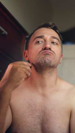 Vertical photo Man uses small trimmer during routine, Gentleman shaves with device in modest washroom atmosphere, Male individual performs grooming task with compact device beforeの写真素材