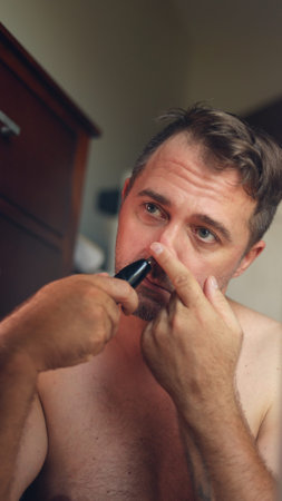 Vertical photo Gentle evening shave, Man quickly grooming in mirror, White man in bathroom carefully shaving face at night, Man with no shirt tending to face using electric razorの写真素材