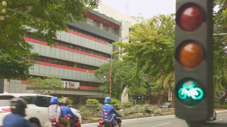 Green bike signal over busy street motorcycle courier maneuvers through traffic past parked cars and delivery scooters, modern office building facade and leafy trees in backgroundのeditorial素材