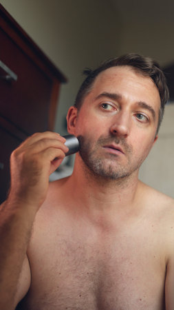 Vertical photo Man grooming indoors, Man attentively grooming face indoors, Individual meticulously caring for facial hair inside home, Man focused on personal grooming inの写真素材