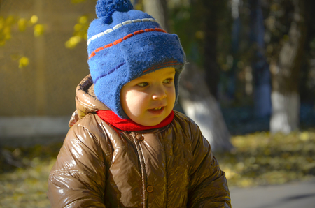 The kid in the autumn park in a blue hat and a brown jacket mysteriously looks away.の写真素材