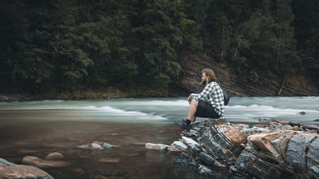A man with a beard in a plaid shirt and shorts sits on the bank of a mountain river, moody colors