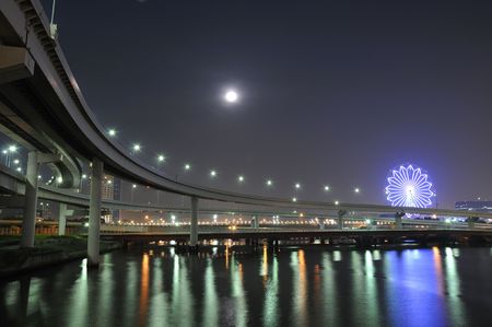  hanged-up highway road over Tokyo bay waters at night time with moon and ferris-wheel illumination on backwardの写真素材