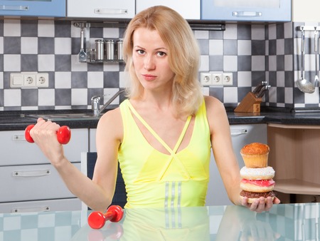Woman choosing between fitness and cakes in the kitchenの写真素材