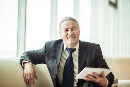 smiling experienced businessman with digital tablet sitting on sofa in the officeの写真素材
