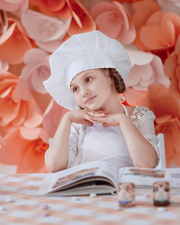 Charming little girl in a chief hat stands at a dinner table ready to cook a dinnerの写真素材