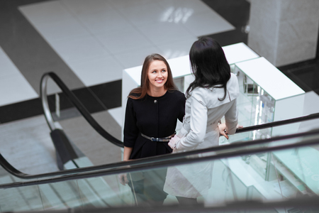 Two smiling business women standing on an escalator in a business centerの写真素材