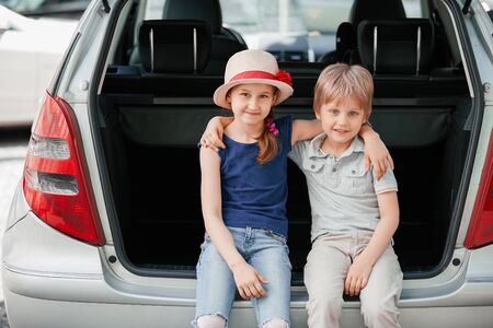 Little sister and brother sitting in the trunk of a car with suitcasesの写真素材