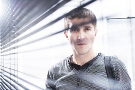 Serious young man looking through window blind