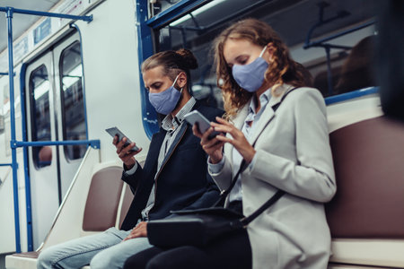passengers wearing protective masks using their smartphones while sitting in a subway car.の写真素材