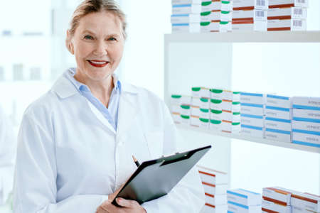 pharmacist with a clipboard and her colleague standing near a pharmacy display case .
