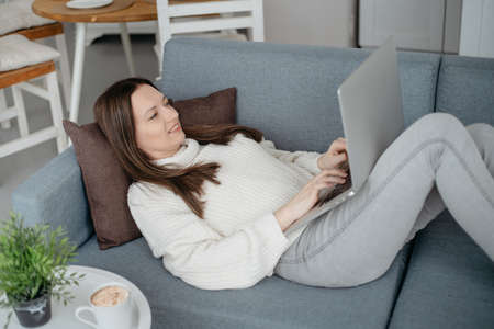smiling young woman with a laptop lying on the sofa in the living room .
