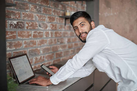 young man using a laptop and a smartphone standing in the kitchen.