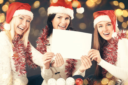 three young girls in costume of Santa Claus showing blank sheets. Beautiful bokeh effect from a garland.の写真素材