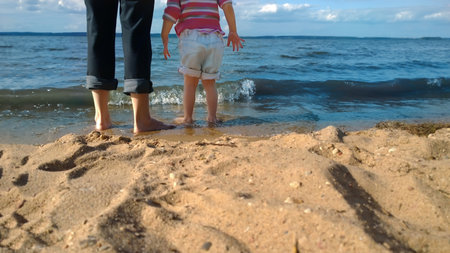 feet of a mother and child stand on the sand by the sea shoreの写真素材