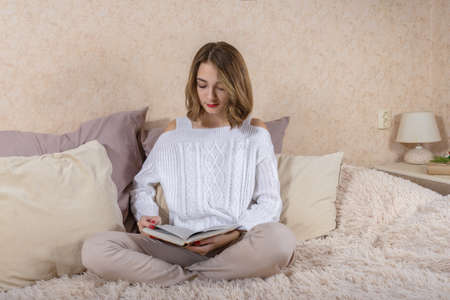 Girl in white sweater is reading a book on the couch