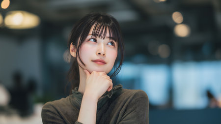 Young woman in thinking pose with hand on chin, blurred office backgroundの素材