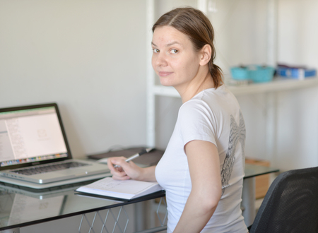 The girl in a white shirt sits at a table with a computer.の写真素材