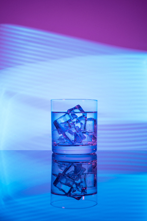 A glass with a drink and ice cubes on a magenta background with blue lights.の写真素材