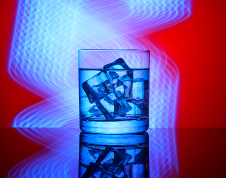 A glass with a drink and ice cubes on a red background with blue lights.の写真素材