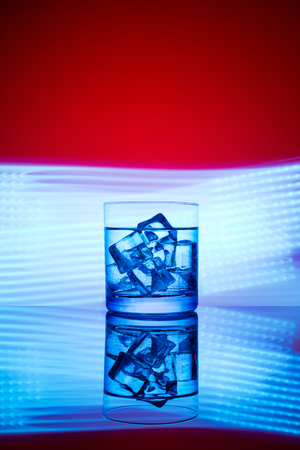 A glass with a drink and ice cubes on a red background with blue lights.の写真素材