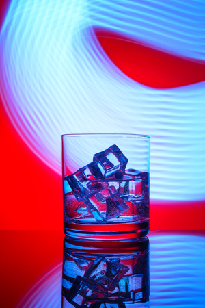 A glass with a drink and ice cubes on a red background with blue lights.の写真素材