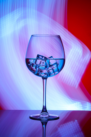 A glass with a drink and ice cubes on a red background with blue lights.の写真素材