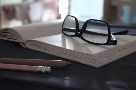 Black glasses on an open book, and two pencils on a dark table.の写真素材
