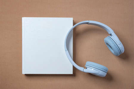 White headphones and a white book on the blue and beige background. Audio book reading online.