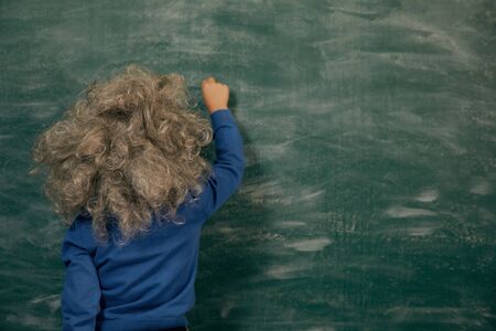 Cheerful smiling little kid (boy) against green chalkboard. Looking at blackboard. School concept