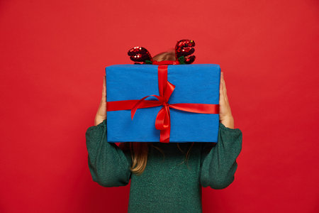 Smiling funny child (kid, girl) in Reindeer Hairband. Holding Christmas gift in hand. christmas concept. Shooting on red backgroundの写真素材