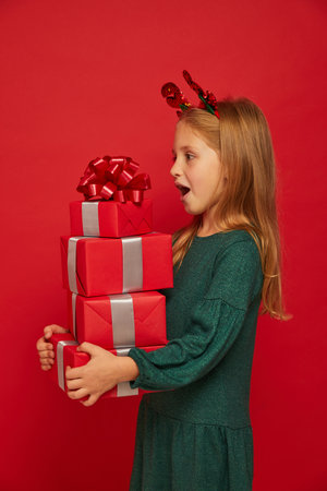 Smiling funny child (kid, girl) in Reindeer Hairband. Holding Christmas gifts in hand. christmas concept. Shooting on red backgroundの写真素材