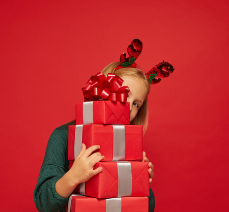 Smiling funny child (kid, girl) in Reindeer Hairband holding Christmas gifts in hand. new year concept. Shooting on red backgroundの写真素材