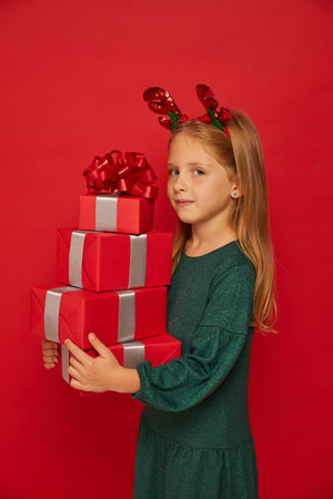 Smiling funny child (kid, girl) in Reindeer Hairband holding Christmas gifts in hand. new year concept. Shooting on red backgroundの写真素材