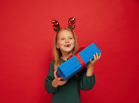Smiling funny child (kid, girl) in Reindeer Hairband holding Christmas gifts in hand. new year concept. Shooting on red backgroundの写真素材