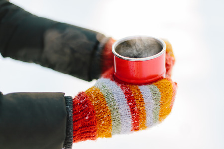 Hands in bright striped mittens holding the red cup outdoors. Soft focus