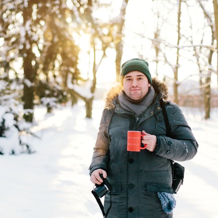 Handsome man standing in a winter forest with cup and cameraの写真素材
