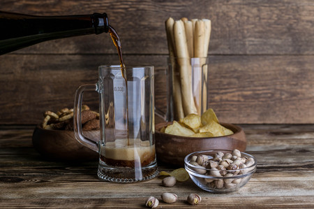 Dark beer from a bottle into a beer mug. Snacks for beer on a wooden table. Chips, croutons, bread sticksの写真素材