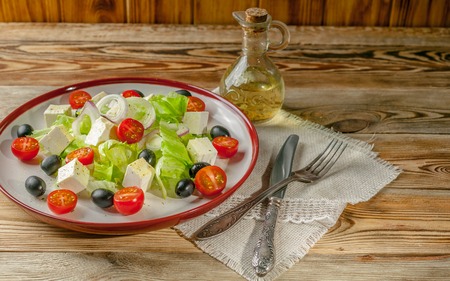 Greek salad with feta cheese, cherry tomatoes and olives in a round plate on a wooden background. Knife and fork on a napkin. Summer salad. Close-upの写真素材