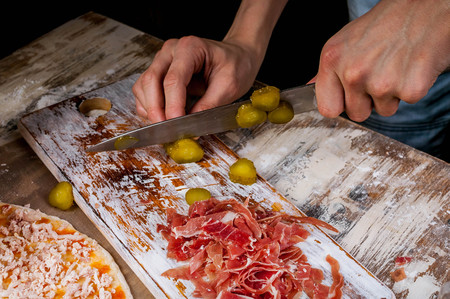 The hands of a young chef cut the vegetables and prosciutto for pizza on a wooden board. Homemade pizza with prosciutto. Close-upの写真素材