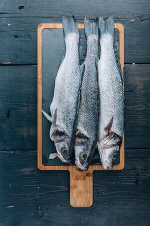 Three sea bass fish on a cutting board on a dark wooden background. Top viewの写真素材