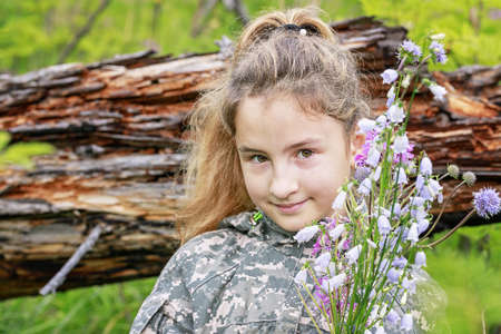 Cute Caucasian girl in camouflage and with a bouquet of forest flowers. Hiking in the forestの写真素材