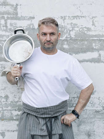 Chef in a white T-shirt with a frying pan in his hands. He has extended arm with thumbs up....