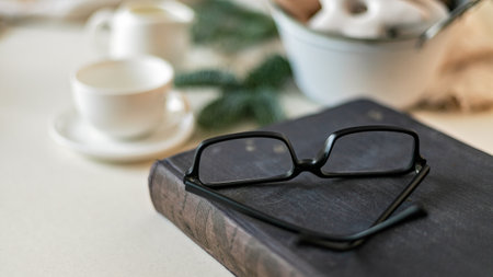 Web banner format. Christmas still life. Old book and glasses in the foreground. White cups and...