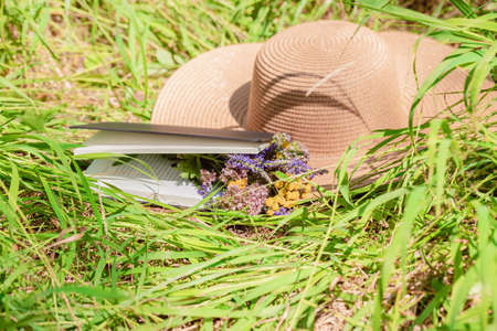 Book, straw hat and a bouquet of forest flowers on the green grass. Summer still lifeの写真素材