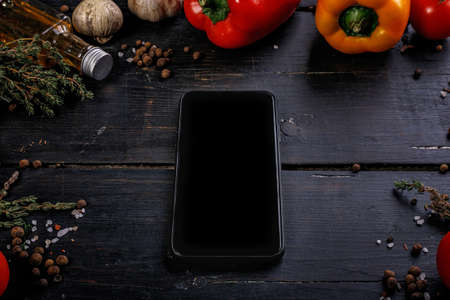 Tomatoes, bell peppers, garlic, olive oil and a cutting board on a black wooden table. Dark contrast food flat lay with vegetables and spices. Black smartphone.の写真素材