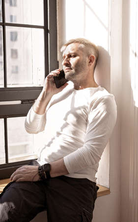 Handsome blonde caucasian man in a white shirt is sitting on a windowsill talking on the phone....
