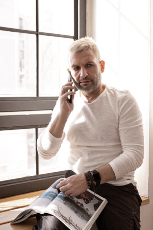 Charming caucasian man in a white shirt is sitting on a windowsill with a journal talking on the...