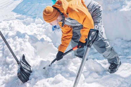 Young caucasian man in a yellow winter suit is holding a snow shovel. A boy is cleaning the backyard...